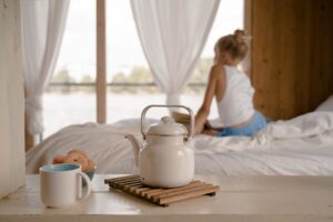 The best teas to help you sleep. A white pot and mug sits on a wooden table, in front of a bed with a woman sitting on it.